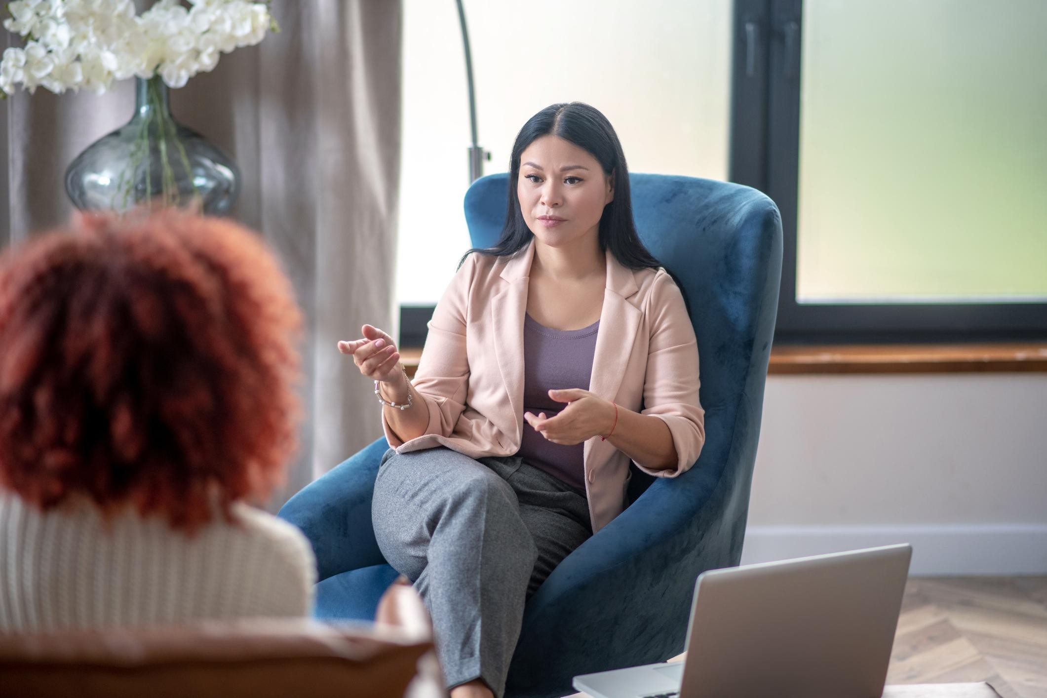 An asian female psychologist sits across from her client and is shown speaking. Next to her, on a little desk, sits her open laptop.