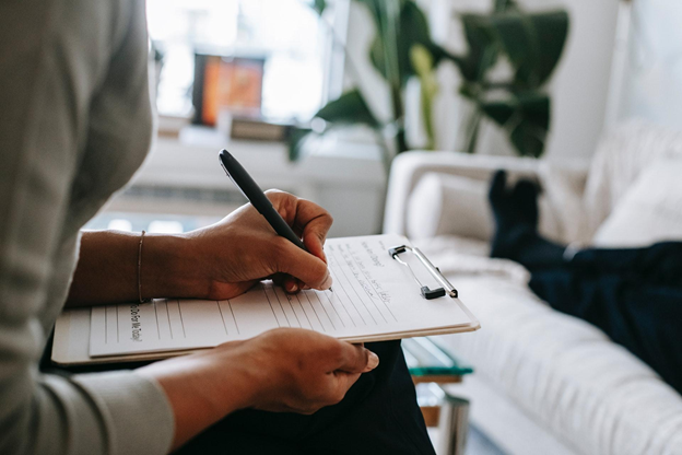 A therapist is seen writing on their clipboard as their client lies on a couch in front of them.