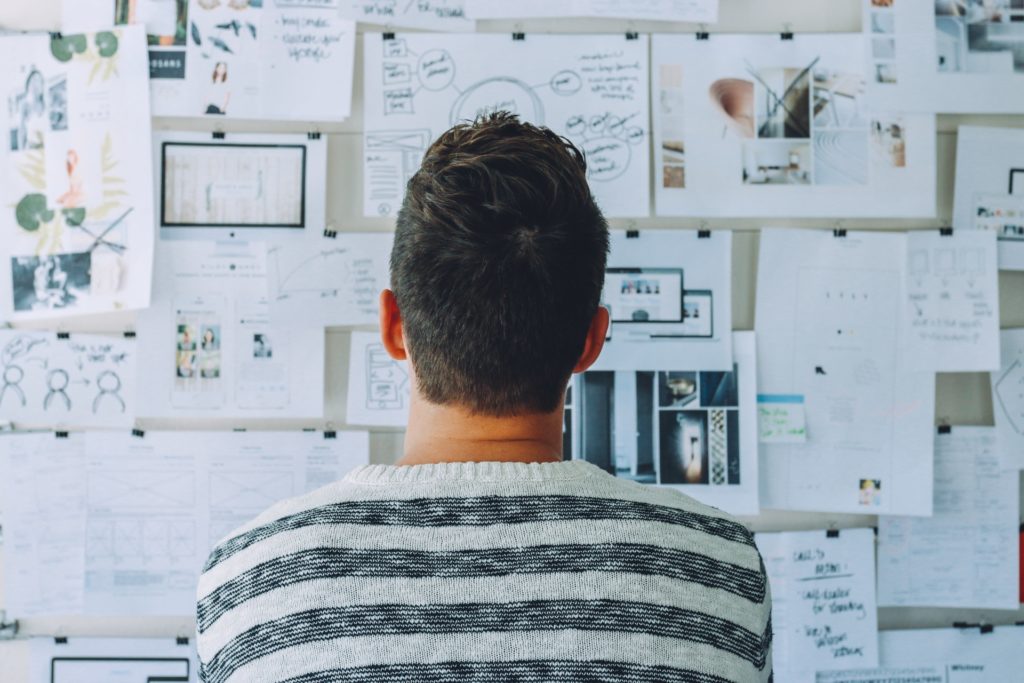 We a see a man in a striped sweater standing in front of a wall covered in papers, drawings, scribbles, and images of all kinds, most likely trying to find a new purpose in his life or figure something out.